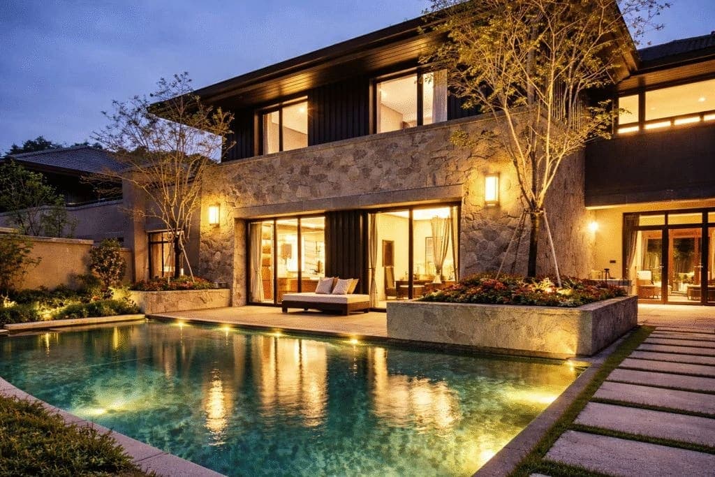 Modern villa at dusk with wall sconces, glowing interiors, uplit trees and a lit swimming pool